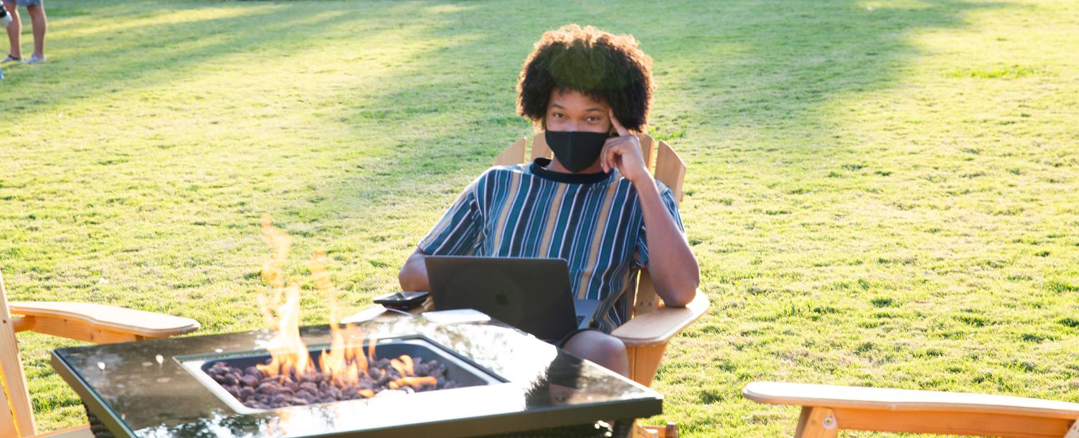 student wearing a mask studying with a laptop sitting by a fire pit