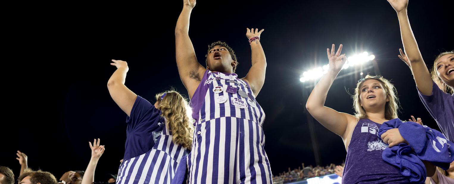 students dressed in purple and white striped overalls, cheering at football game