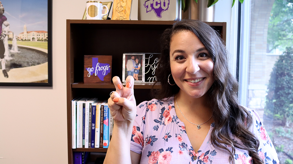 Dark haired woman standing in front of a bookcase, wearing a floral purple and pink blouse, giving horned frogs hand sign