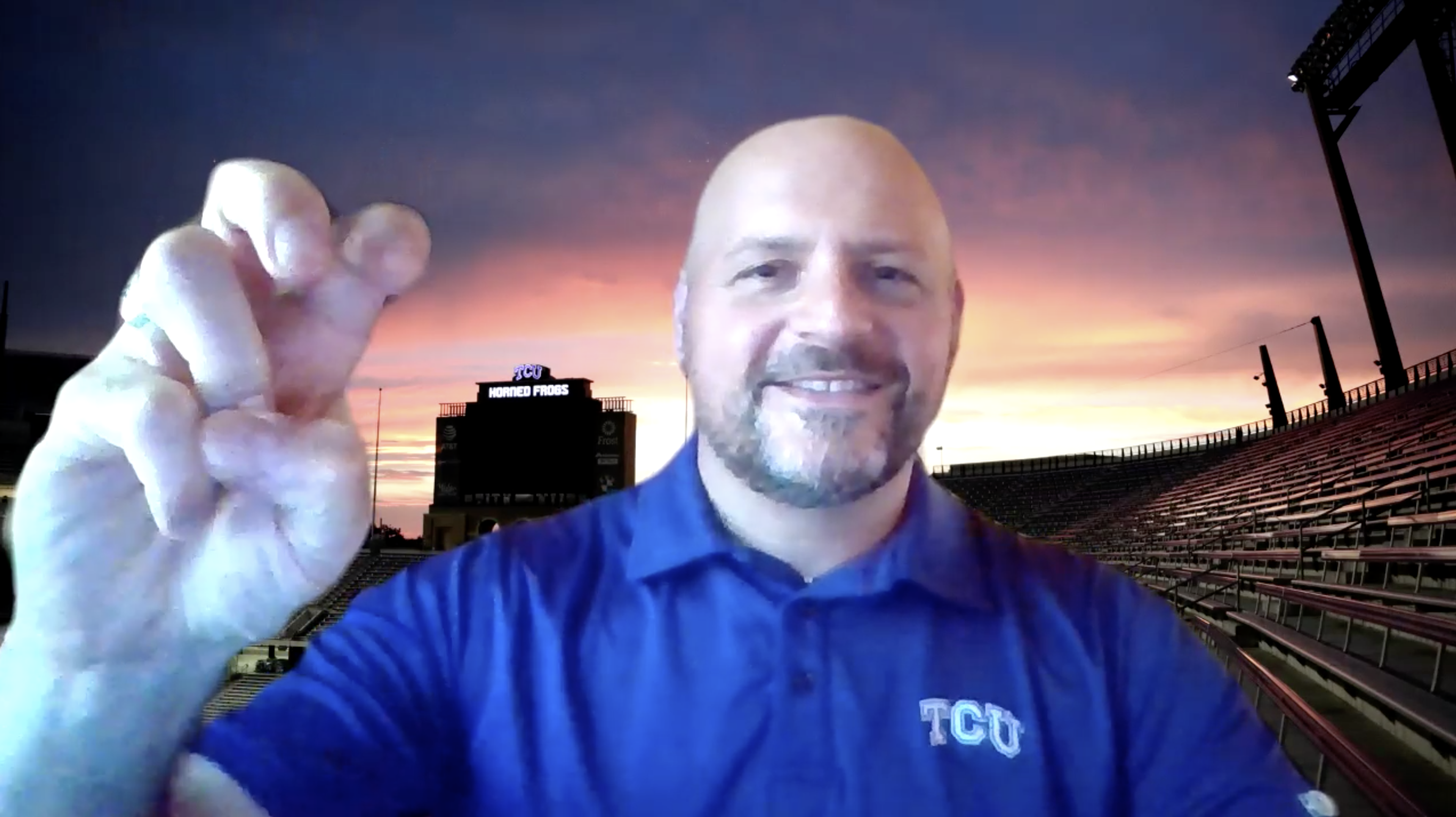 Bald man wearing purple TCU shirt and giving the horned frog hand sign. Background shows sunset at football stadium